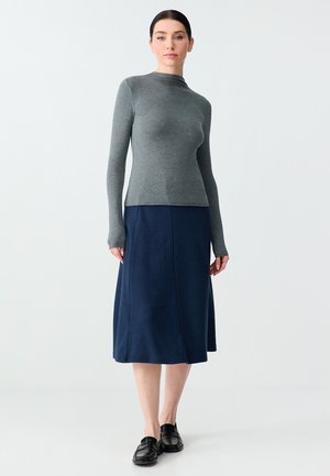 Kobieta stojąca, ubrana w dopasowaną szarą bluzkę z długim rękawem, granatową spódnicę midi i czarne mokasyny, na tle jasnego, jednolitego tła.