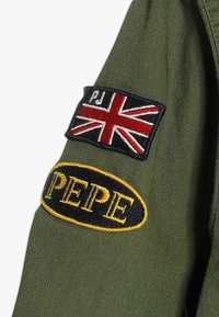 Zielona rękawica kurtki z tkaniny z naszywką brytyjskiej flagi oraz czarną owalną odznaką "PEPE" z żółtym haftem.