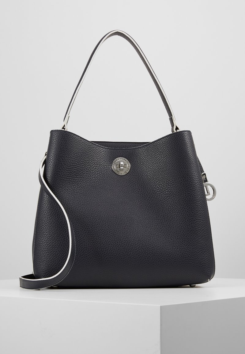 Sac à main en cuir noir texturé avec fermoir à tourniquet argenté, une poignée supérieure unique et une bandoulière amovible, posé sur une surface blanche.