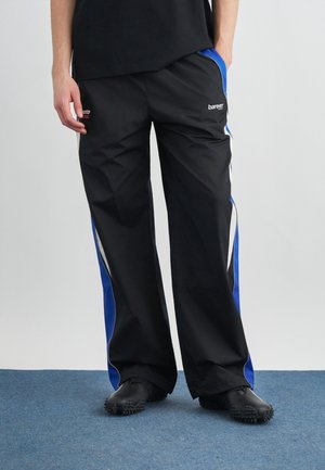 Άτομο που φοράει μαύρο παντελόνι φόρμας με μπλε και λευκές ρίγες στο πλάι και μαύρα παπούτσια με υφή στην σόλα, στέκεται σε μπλε χαλί.