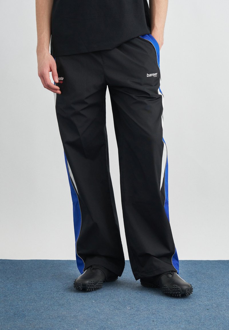 Άτομο που φοράει μαύρο παντελόνι φόρμας με μπλε και λευκές ρίγες στο πλάι και μαύρα παπούτσια με υφή στην σόλα, στέκεται σε μπλε χαλί.