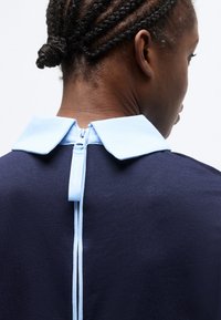 Άτομο με πλεξούδες, που φαίνεται από πίσω, φοράει ένδυμα ναυτικού χρώματος με ανοιχτό μπλε γιακά και κεντρικό φερμουάρ στο πίσω μέρος.