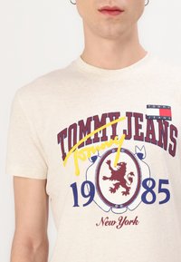 Krēmkrāsas kokvilnas T-krekls ar drukātu logotipu ar bordo un zilu akcentiem, lauvas emblēmu un dzeltenu izcelšanu uz zīmola nosaukuma.