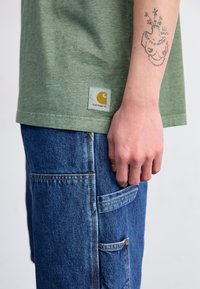 Grønn bomulls t-skjorte med et Carhartt-logo-patch, kombinert med blå denim cargo-bukser med flere lommer og gule sømdetaljer.