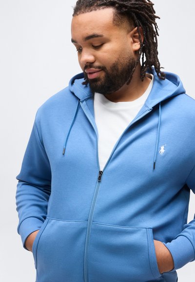 Άνδρας με γένια και dreadlocks, φοράει μπλε φούτερ με φερμουάρ και τσέπες και λευκό μπλουζάκι, κοιτάζοντας προς τα κάτω αριστερά του.