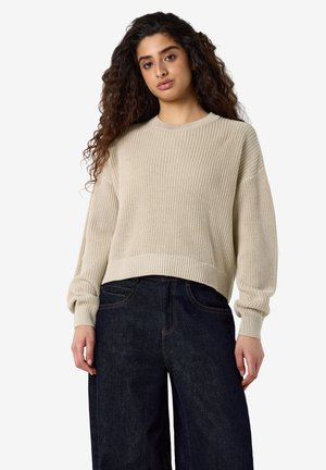 Jeune femme aux longs cheveux bouclés portant un pull en tricot beige et un jean large taille haute foncé, debout devant un fond uni.
