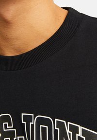 Black sweatshirt fabric with a ribbed round neckline and bold white lettering in a stylized font across the chest area.