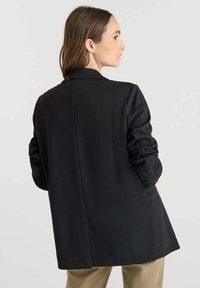 Blazer noir au design cintré, doté d'une fente unique dans le dos, de manches longues et d'une texture lisse. Sans matériel ou motifs visibles.