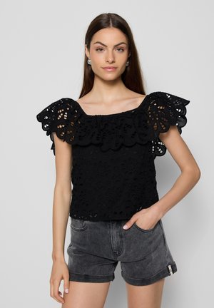 Zwarte katoenen blouse met geborduurde uitsnijdingen en flare mouwen. Vierkante halslijn; gecombineerd met grijze hoge shorts.
