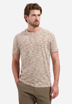 Mann mit kurzen lockigen Haaren und Bart, der ein beige-braun gestreiftes kurzärmliges Hemd und olivgrüne Hosen trägt, steht mit einer Hand in der Tasche.