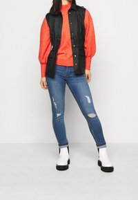 Svart ärmlös väst med knappstängning, röd stickad tröja, slitna blå jeans och vita ankelstövlar med svarta accenter.