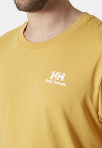 Camiseta de algodón amarilla con cuello redondo. Presenta el logo blanco "HH" y el texto "Helly-Hansen" en el lado izquierdo del pecho. Textura suave. Diseño minimalista.