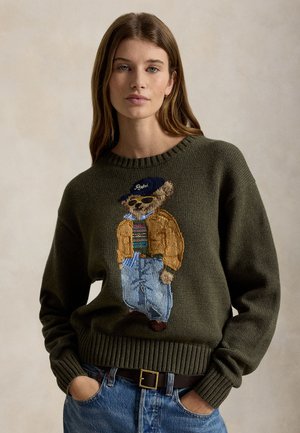 Femme portant un pull vert olive avec un ours brodé habillé d'une veste décontractée, une chemise rayée, un jean, des lunettes de soleil et une casquette, debout les mains dans les poches.