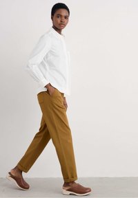Camicia bianca con bottoni e maniche lunghe, pantaloni giallo-marroni e zoccoli marroni. Tessuto morbido, vestibilità rilassata, con pantaloni alla caviglia e polsini arrotolati.