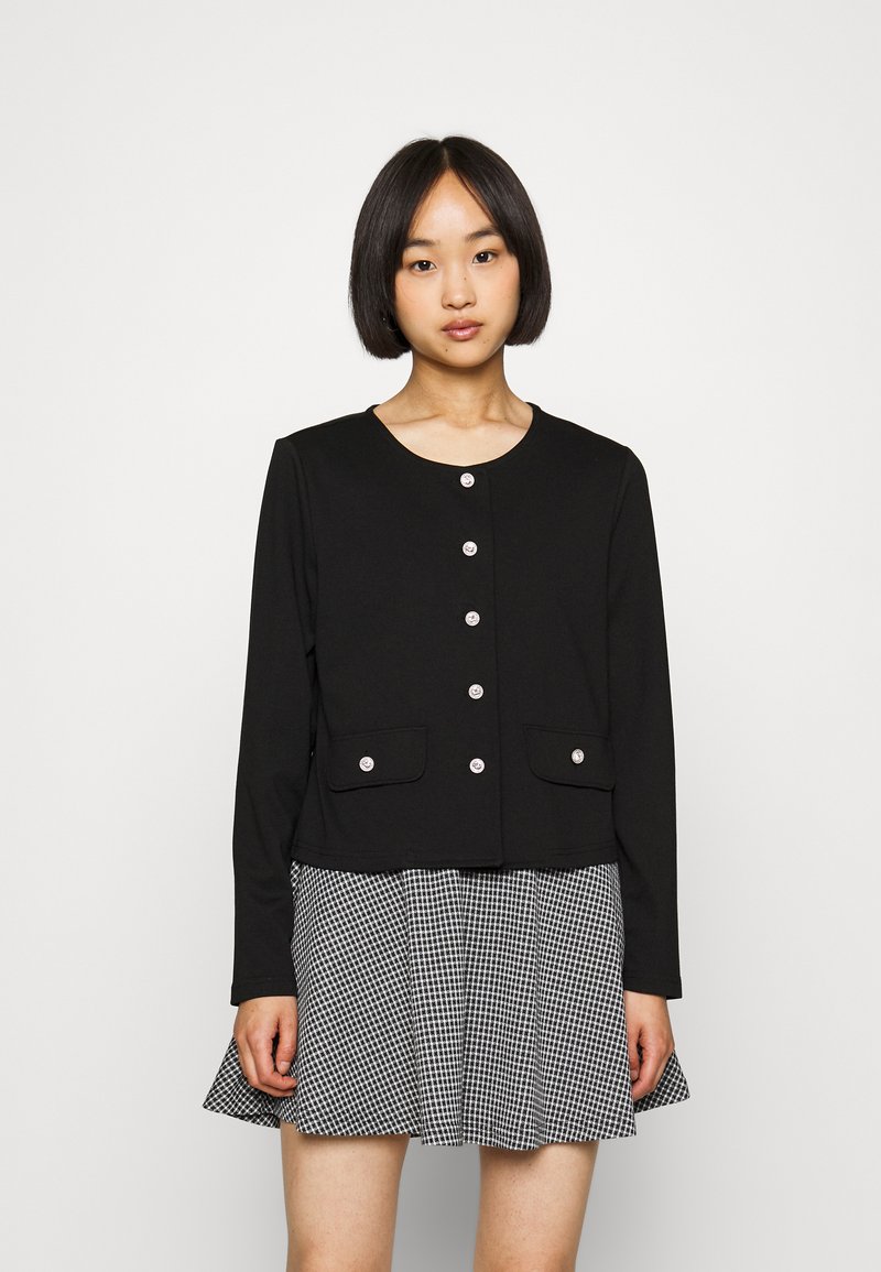 Fekete, sima anyagú, rövid kabát hosszú ujjal, elülső gombokkal és két zsebbel. Fekete-fehér kockás miniszoknyával párosítva.