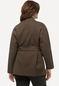 Giacca marrone trapuntata con design avvolgente, maniche ampie e vita con cintura. Presenta una texture a motivo romboidale e una vestibilità rilassata.