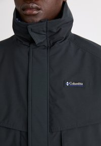 Schwarze Columbia-Jacke mit hohem Kragen, Frontreißverschluss und Logo-Patch auf der linken Brust. Glattes, wasserabweisendes Material.