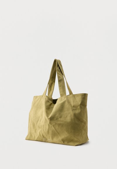 Grand sac fourre-tout en toile marron clair avec doubles anses, tissu légèrement froissé et texte imprimé discret près du bord inférieur.