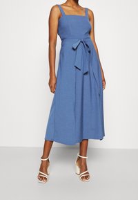 Mujer vistiendo un vestido midi sin mangas de color azul con escote cuadrado y cintura anudada, combinado con sandalias de tacón bloque blancas con tiras.