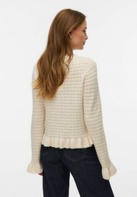 Krämvit stickad tröja med texturerat mönster, ribbad nederkant och utsvängda ärmar. Bärs med mörka jeans.