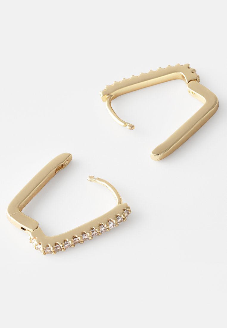 Boucles d'oreilles créoles en ton or avec une forme géométrique, ornées d'une rangée de pierres claires le long du bord inférieur et d'une finition lisse et polie.