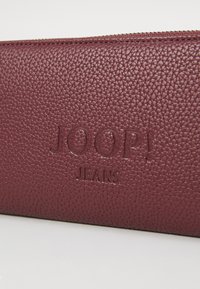 Burgundska usnjena denarnica s teksturirano površino, z embosiranim logotipom "JOOP! JEANS" in zadrgo ob zgornjem robu.