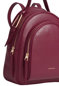 Bordeaux lederen rugzak met een gladde textuur, ronde vorm, gouden hardware accenten en dubbele ritsen op de voorcompartimenten. Gouden logo weergegeven.