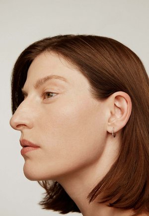 Perfil de una mujer con cabello corto y castaño, piel clara, que lleva un pequeño pendiente de aro plateado, sobre un fondo claro y sencillo.