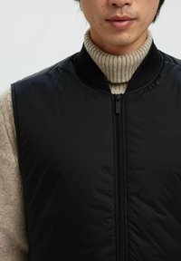 Crna pufasta prsluk s visokim ovratnikom, s prednjim patentnim zatvaračem i rebrastim detaljima. Nosi se preko bež, teksturirane dolčevite.