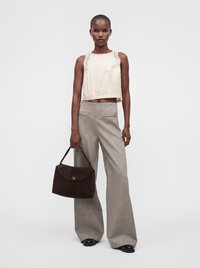 Personne debout porte un haut crème sans manches avec bordure en dentelle, un pantalon large gris, des chaussures noires et tient un grand sac à main marron d'une main.