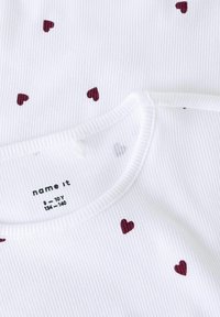 Camiseta blanca de canalé con pequeños patrones de corazones color marrón, con un escote redondo y detalle de botones en la parte posterior. Etiqueta de tamaño visible.
