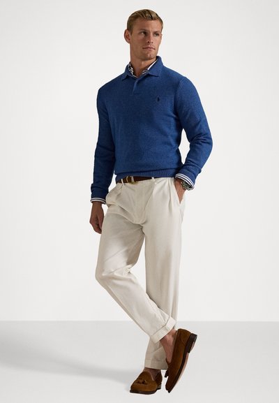 Blå strikket sweater med polo krave og ribbestrikkede manchetter, parret med cremefarvede bukser og brune loafers. Modellen står foran en neutral baggrund.