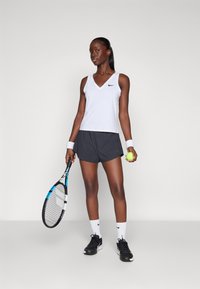 Conjunto deportivo que incluye una camiseta sin mangas de color gris claro y pantalones cortos negros. Los accesorios incluyen una raqueta de tenis, una pelota y muñequeras blancas.