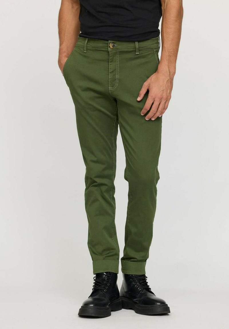 Chinos en coton verts avec une coupe slim, présentant une fermeture à bouton, des passants de ceinture et des manches retroussées, associés à des bottes noires à lacets.
