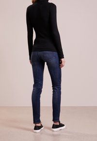 Schwarz langärmliges Rollkragenoberteil, dunkelblaue enge Jeans und schwarze Sneakers, von hinten gegen einen hellen Hintergrund betrachtet.