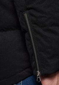 Czarna kurtka z zamkiem błyskawicznym na mankietach. Materiał jest gładki z matowym wykończeniem. Posiada metalowy zamek błyskawiczny i wzmocnione szwy wzdłuż szwów.