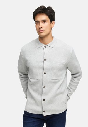 Hombre vistiendo un cárdigan de botones gris claro con dos bolsillos frontales, combinado con jeans azul oscuro, de pie contra un fondo blanco.