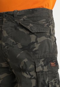 Pantaloni cargo mimetici in verde scuro e nero, con chiusura a pulsante, tasche laterali e un'etichetta tessuta che dettagli il marchio.