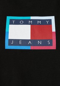 Czarny materiał z prostokątnym logo zawierającym czerwone, białe, niebieskie i morski elementy. Napis "TOMMY JEANS" dużymi białymi literami.