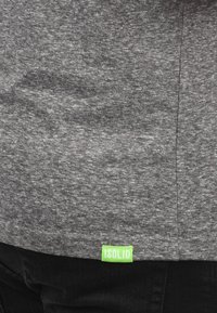 Camiseta de tela texturizada gris con un ligero patrón jaspeado, que presenta una pequeña etiqueta verde de iSOLID en el dobladillo. Pantalones vaqueros negros visibles por debajo.