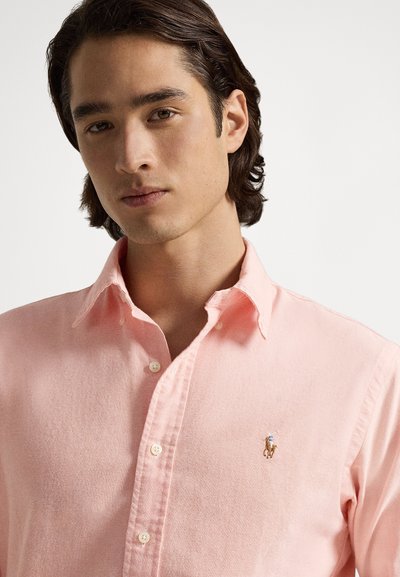 Svetlo roza srajca s klasičnim ovratnikom, z majhnim vezenim logotipom konja na levi strani prsi in teksturiranim detajlom tkanine.