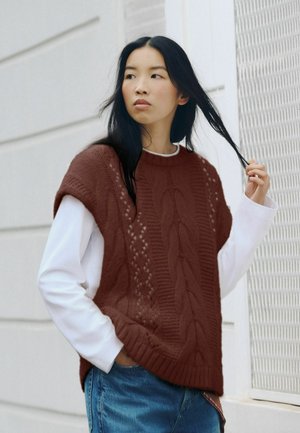 Donna con lunghi capelli neri che indossa un gilet di maglia marrone sopra una camicia bianca a maniche lunghe e jeans blu, in piedi accanto a un muro bianco, toccandosi i capelli.