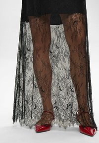 Melnas mežģīņu pārklājs ar ziedu rakstiem un caurspīdīgām daļām, atklājot ādas toni zivju tīkla zeķes. Sarkanās norādītās kurpes uzsver šo izskatu.