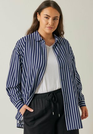 Camisa blanca y azul marino a rayas, de botones y corte relajado, con un bolsillo en el pecho y mangas largas, llevada sobre una camiseta blanca.