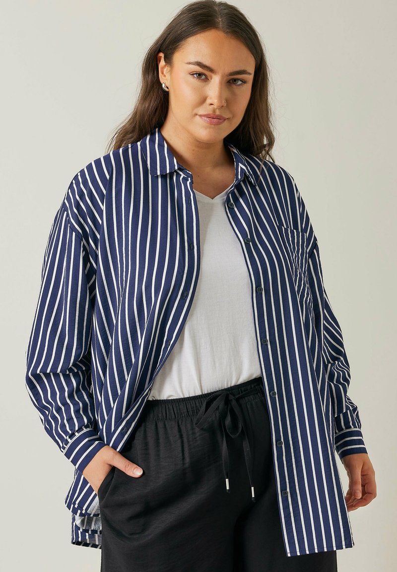Camisa blanca y azul marino a rayas, de botones y corte relajado, con un bolsillo en el pecho y mangas largas, llevada sobre una camiseta blanca.