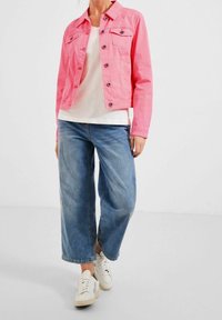 Woman wearing a bright pink buttoned jacket, white top, wide-leg blue jeans, and white sneakers standing against a plain background.