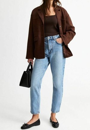 Kvinde iført brun knapjakke, sort top, lyseblå jeans, sorte flade sko, holder sort håndtaske med den ene hånd i lommen.