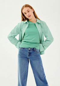 Młoda kobieta z długimi blond włosami, ubrana w zielony prążkowany top, zielono-biały w paski T-shirt i niebieskie dżinsy, z rękami w kieszeniach.
