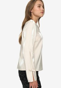 Shiny ivory long-sleeve blouse with side slits and button accents. Smooth texture. Paired with black sequined pants.