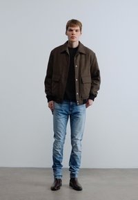 Καφέ υφασμάτινο σακάκι με δύο μπροστινές τσέπες, μαύρο τοπ από κάτω, ανοιχτό μπλε τζιν με σκισίματα και σκούρα καφέ δερμάτινα μποτάκια.